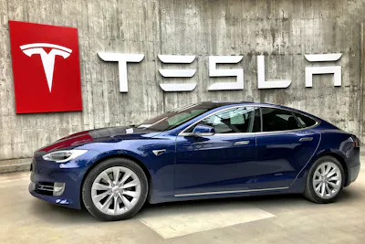 The image shows a Tesla Model S car parked in front of a large Tesla logo, with a concrete wall background.